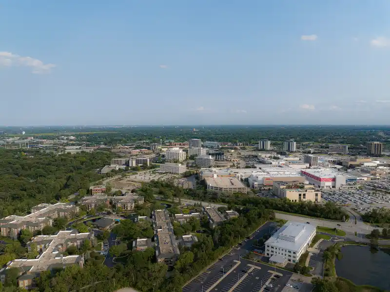 Aerial view of a suburban area with buildings, parking lots, and greenery under a clear sky.