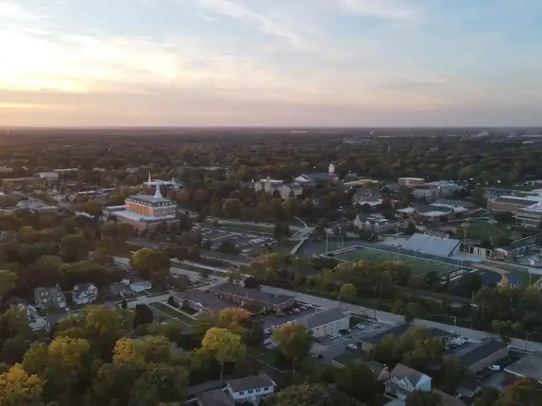 Aerial view of a suburban area with a large building, sports field, and many trees at sunset.