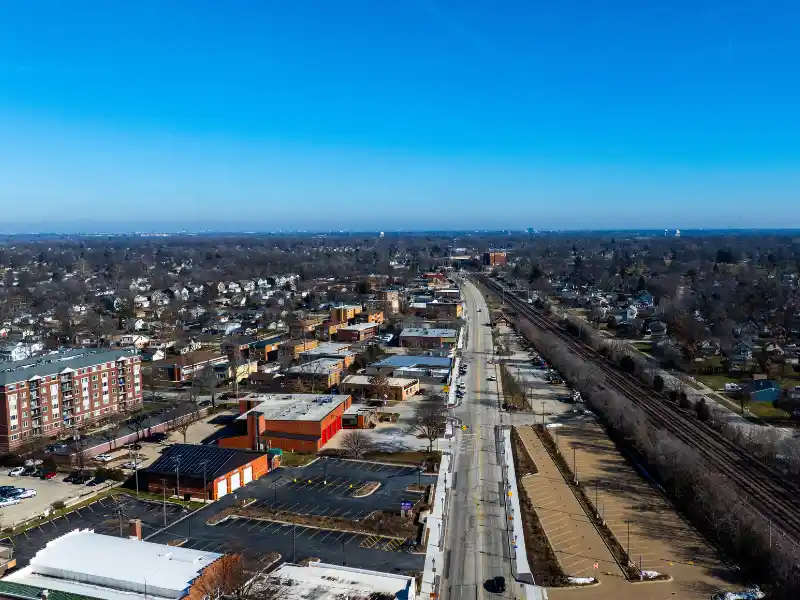 Aerial view of a suburban area with a main road, buildings, houses, and a railway track under a clear blue sky.
