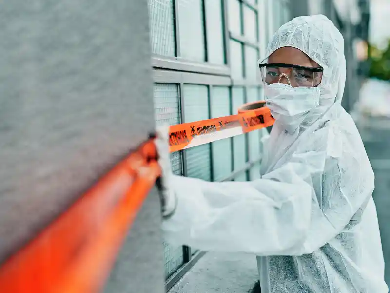 Person in full protective suit and mask placing orange caution tape on a building exterior.