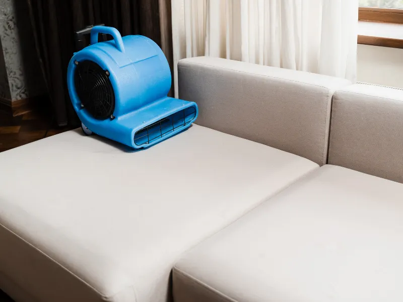 Blue air blower placed on a beige sofa near a window with white curtains.