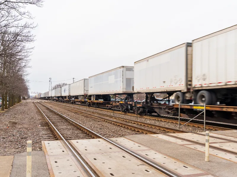 Long freight train carrying white cargo containers on railway tracks beside leafless trees.
