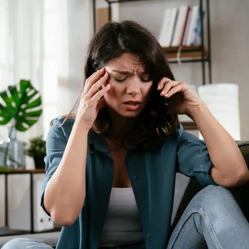Woman sitting on a couch holding a phone to her ear and touching her temple with a pained expression.