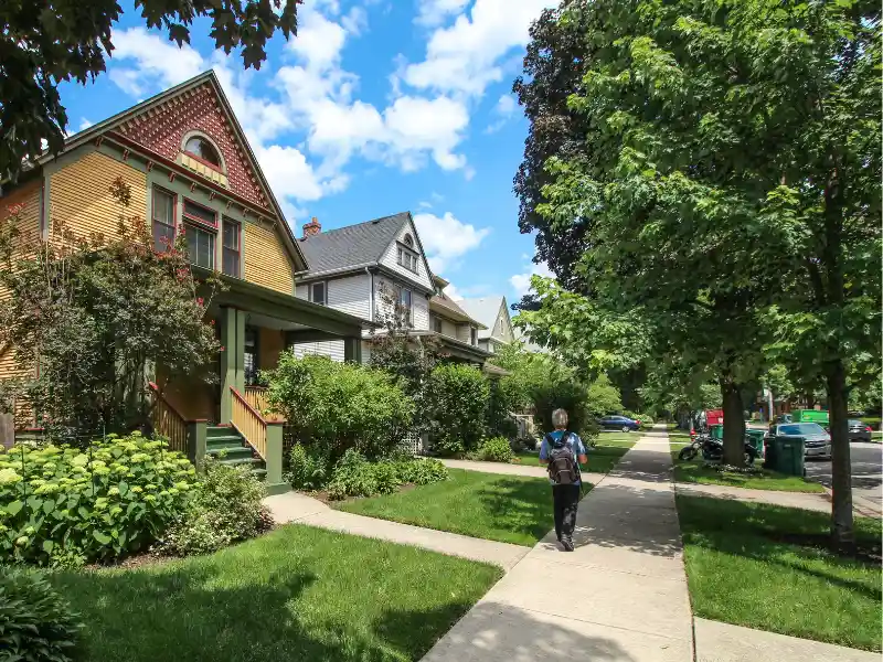 Person walking on a sidewalk past a row of houses with green lawns and trees on a sunny day.
